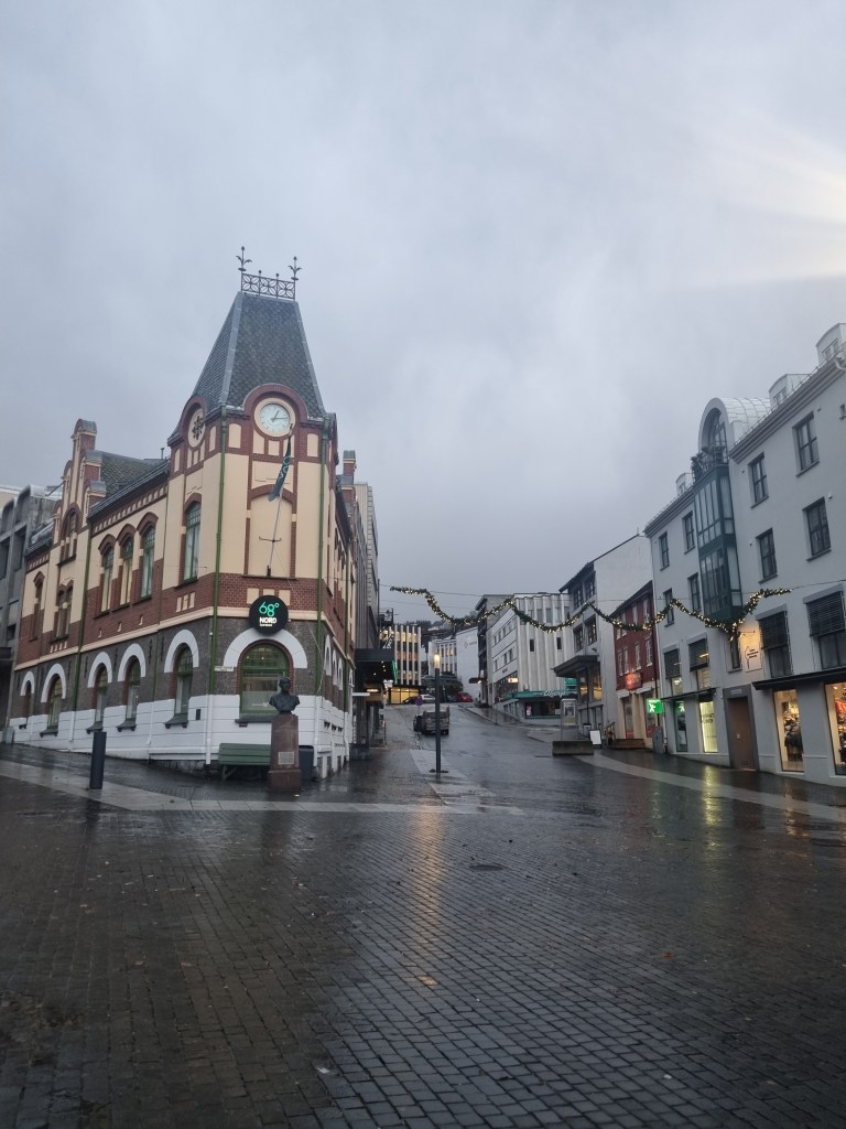 Eine verregnete Stadtstraße mit historischen Gebäuden, einschließlich eines auffälligen Uhrturms, in einer grauen Umgebung.