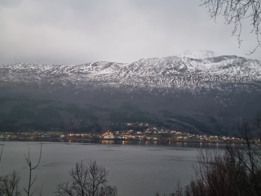 Blick auf einen schneebedeckten Berg im Hintergrund, mit einem ruhigen See und einem kleinen, beleuchteten Dorf im Vordergrund. Der Himmel ist bewölkt, was eine gedämpfte Lichtstimmung erzeugt.